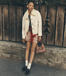 A woman wearing a white shearling jacket and a patterned skirt stands in front of a rustic wooden fence and stone wall. carousel Product Image 1