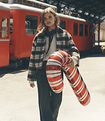 A woman wearing a plaid jacket and jeans holds a large candy cane balloon in front of a red train. carousel Product Image 1