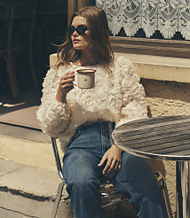 A woman wearing sunglasses and a textured white blouse sits at a small outdoor table with a cup of coffee, positioned against a rustic building wall and window. carousel Product Image 1