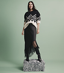 A smiling woman wearing a patterned sweater, black skirt, and boots stands on a silver foil pedestal against a solid light green background. carousel Product Image 4