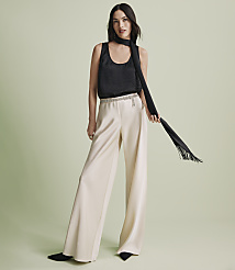 A woman wearing a black sleeveless top, wide-leg cream trousers, and a long black scarf stands against a plain light green background. carousel Product Image 4