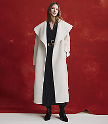 A woman wearing a long white coat over a black pleated dress and black boots stands against a textured red background. carousel Product Image 5