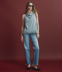 A woman wearing a light blue cowl-neck top with fringe, blue jeans, and black heels stands against a solid red background. carousel Product Image 4