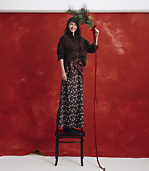 A smiling woman wearing a brown fringed cardigan and a floral skirt stands on a wooden chair against a textured red backdrop while holding a pine branch. carousel Product Image 5