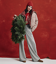 A woman wearing a red beanie, a checkered jacket, and wide-leg grey trousers holds a large evergreen wreath against a textured red background. carousel Product Image 4