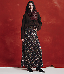 A woman wearing a brown cardigan, a red patterned scarf, and a long floral skirt stands against a textured red background. carousel Product Image 4