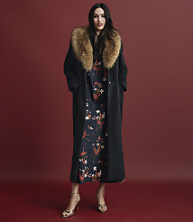 A woman wearing a long black coat with a fur collar over a floral dress and gold heels stands against a solid red background. carousel Product Image 5