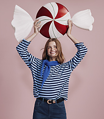 A smiling woman in a blue striped shirt holds a giant red and white peppermint candy against a solid pink background. carousel Product Image 4