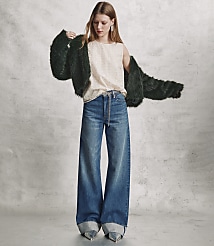 A woman wearing a dark green faux fur coat, a cream top, and wide-leg blue jeans stands against a textured grey concrete wall. carousel Product Image 4