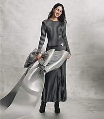 A smiling woman in a long, ribbed grey dress holds a large piece of shimmering silver fabric against a plain, textured grey concrete wall. carousel Product Image 4