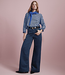 A woman wearing a blue and white striped shirt, a blue neck scarf, and wide-leg blue trousers stands against a solid pink background. carousel Product Image 3