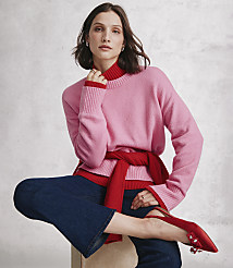 A woman wearing a pink sweater, red turtleneck, and blue jeans sits against a neutral, textured grey wall. carousel Product Image 2