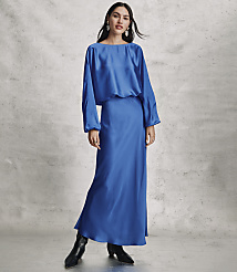 A woman wearing a long, royal blue satin dress and black boots stands against a textured grey concrete wall. carousel Product Image 2