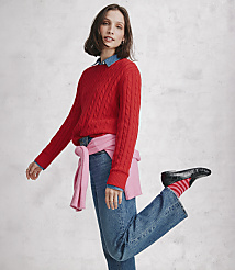 A woman wearing a red cable-knit sweater and blue jeans poses against a neutral, textured grey wall. carousel Product Image 4