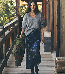 A woman wearing a grey sweater and a navy sequined skirt holds a holiday wreath while walking along a rustic wooden porch. carousel Product Image 1