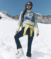 A smiling woman in a grey sweater and blue leggings stands on a snowy field with rugged mountains under a clear blue sky in the background. carousel Product Image 1