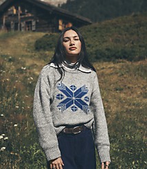 A woman wearing a grey patterned sweater and blue trousers stands in a grassy field with a rustic wooden cabin in the background. carousel Product Image 1