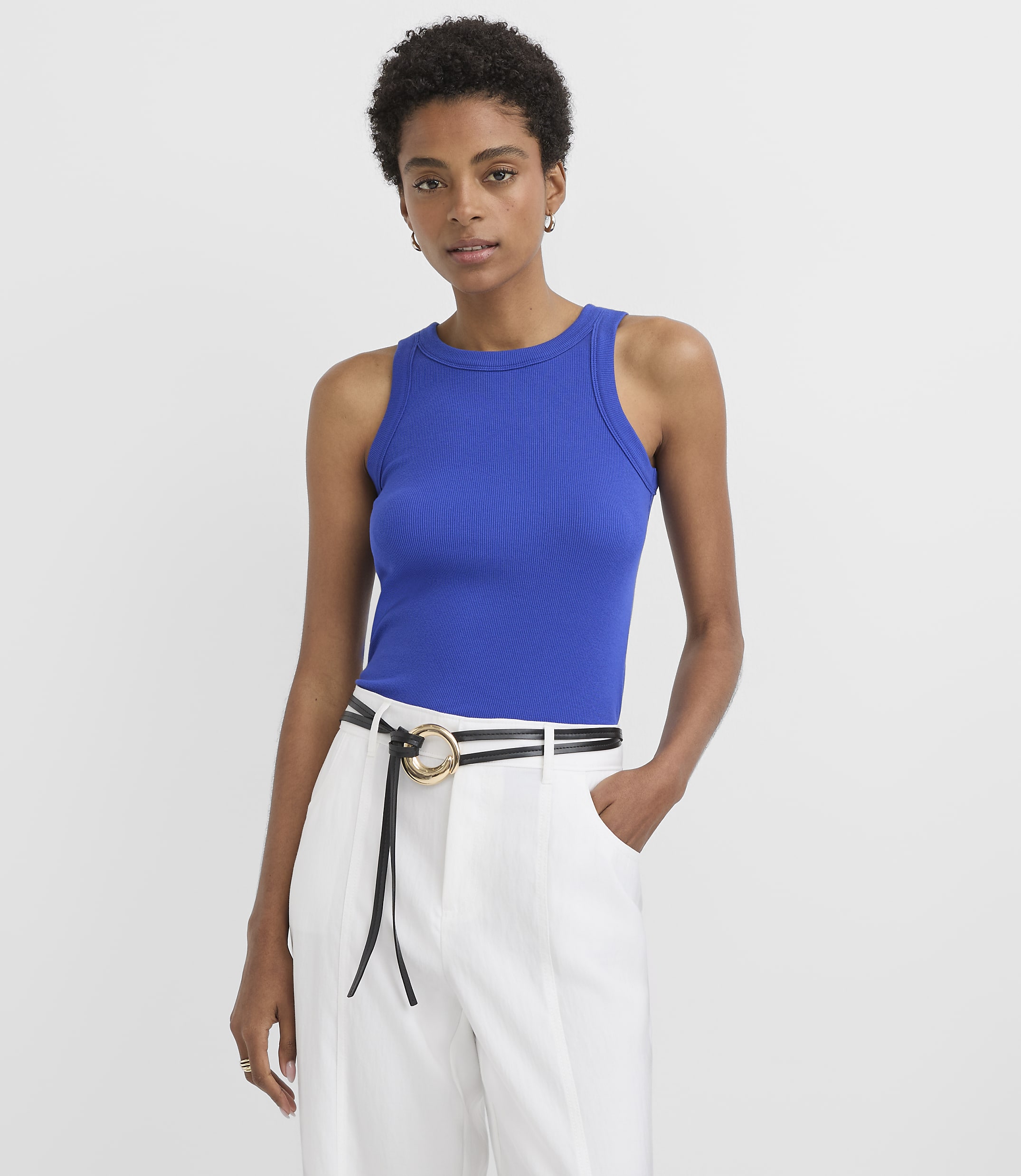 A woman in a blue ribbed tank top and white pinstripe pants stands against a plain white background.