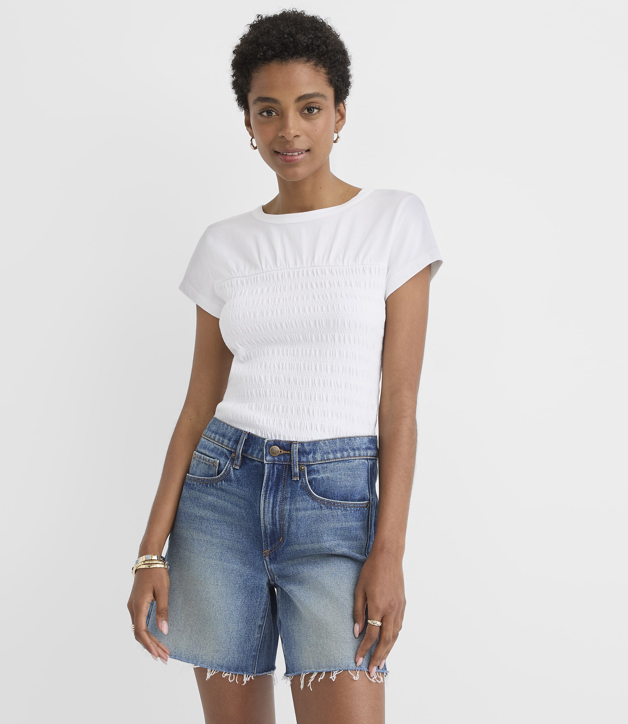 A woman in a white smocked t-shirt and denim shorts stands against a plain white background.