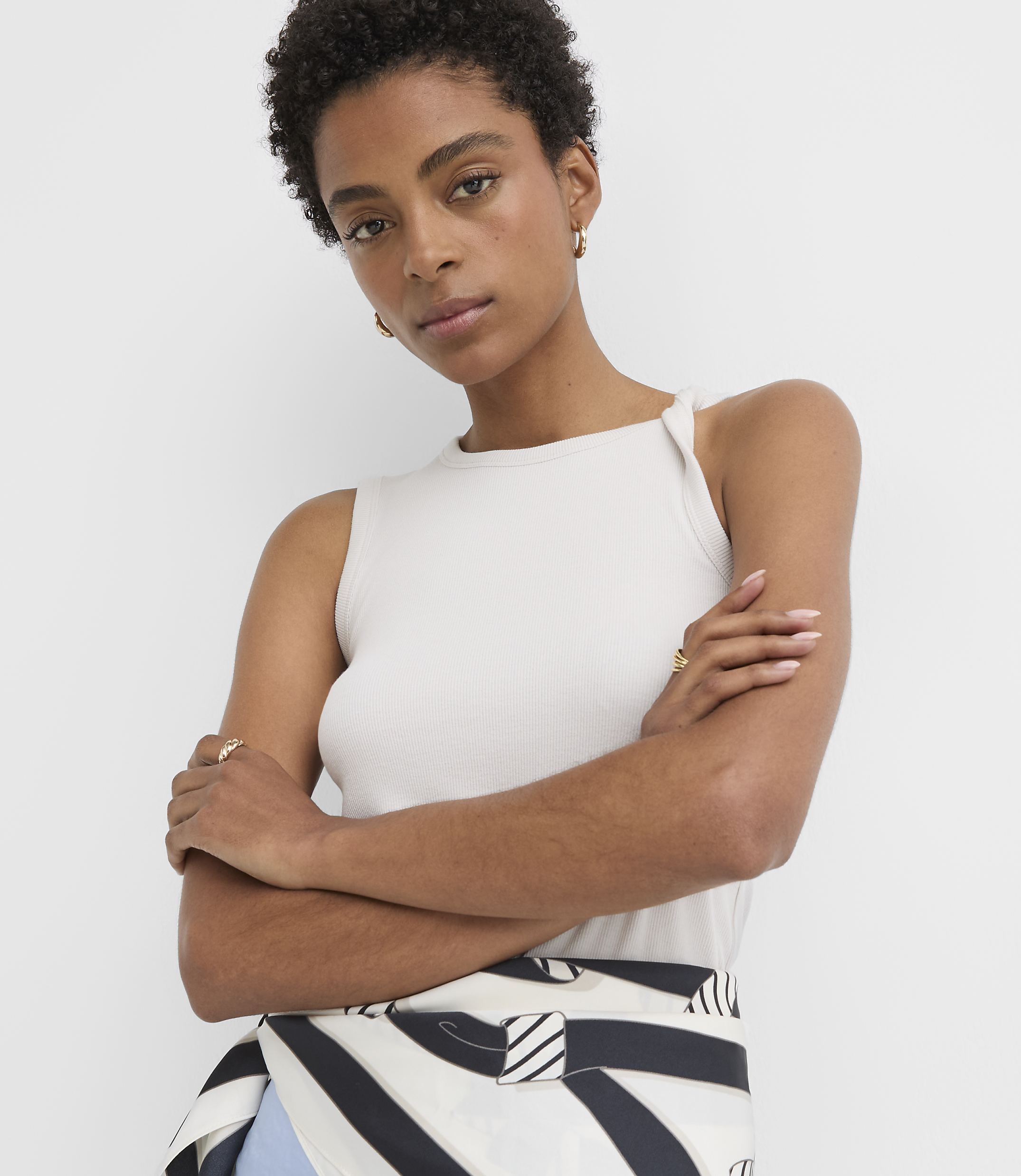 A woman with short dark hair and a light-colored tank top poses with her arms crossed in front of a plain white background.