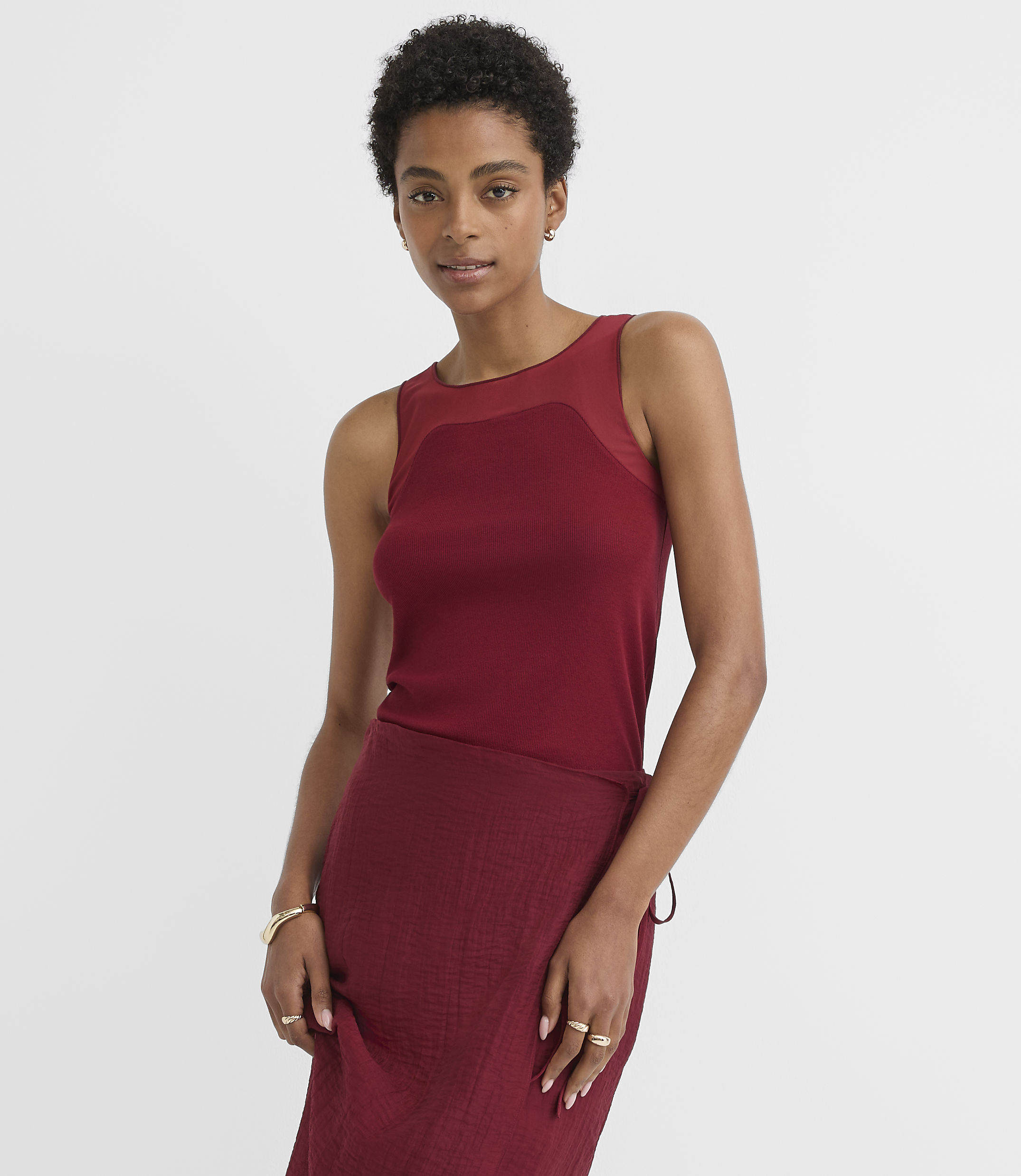 A woman in a maroon ribbed tank top and skirt stands against a plain white background.