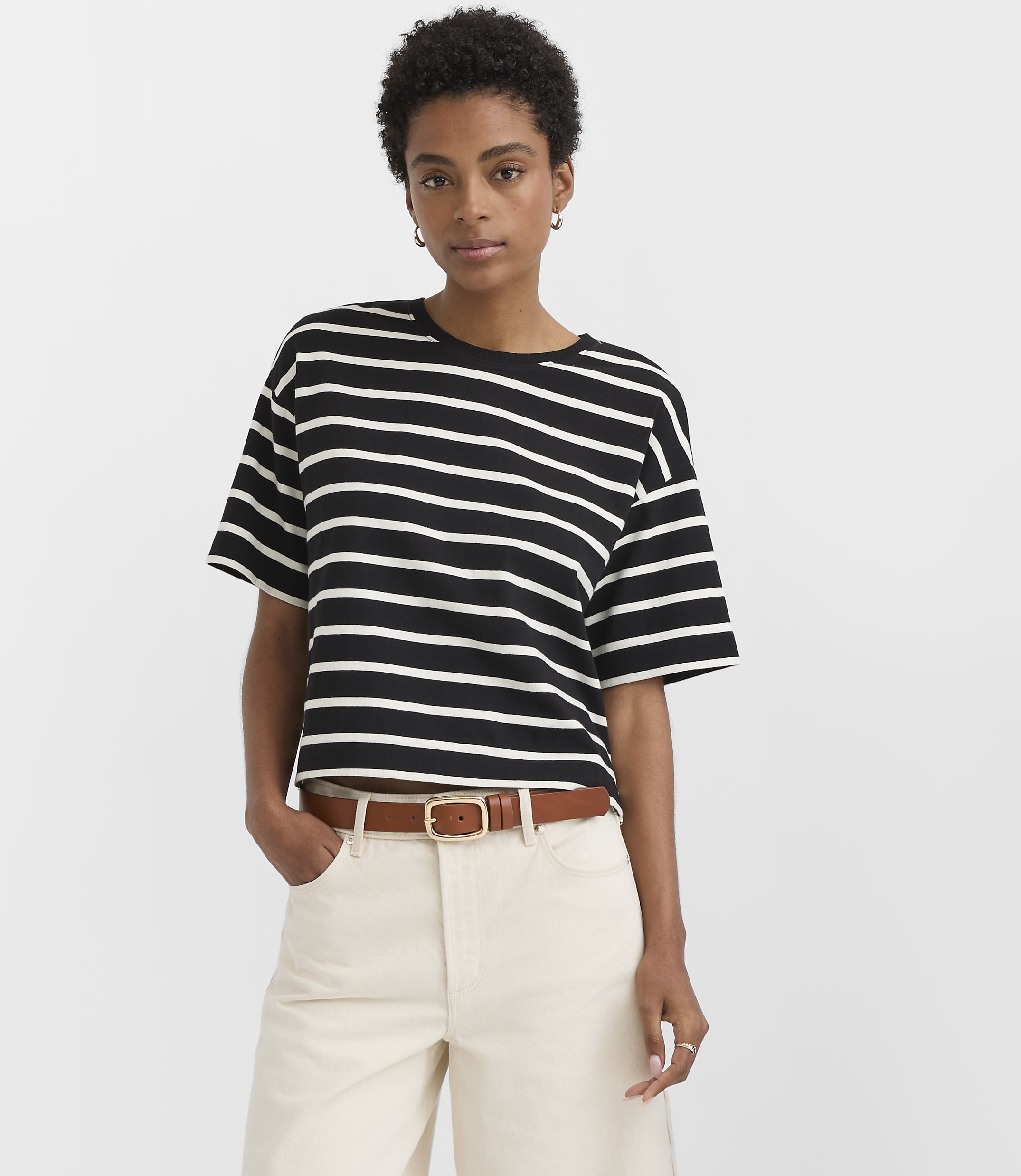 A woman in a black and white striped t-shirt and cream pants stands against a plain white background.