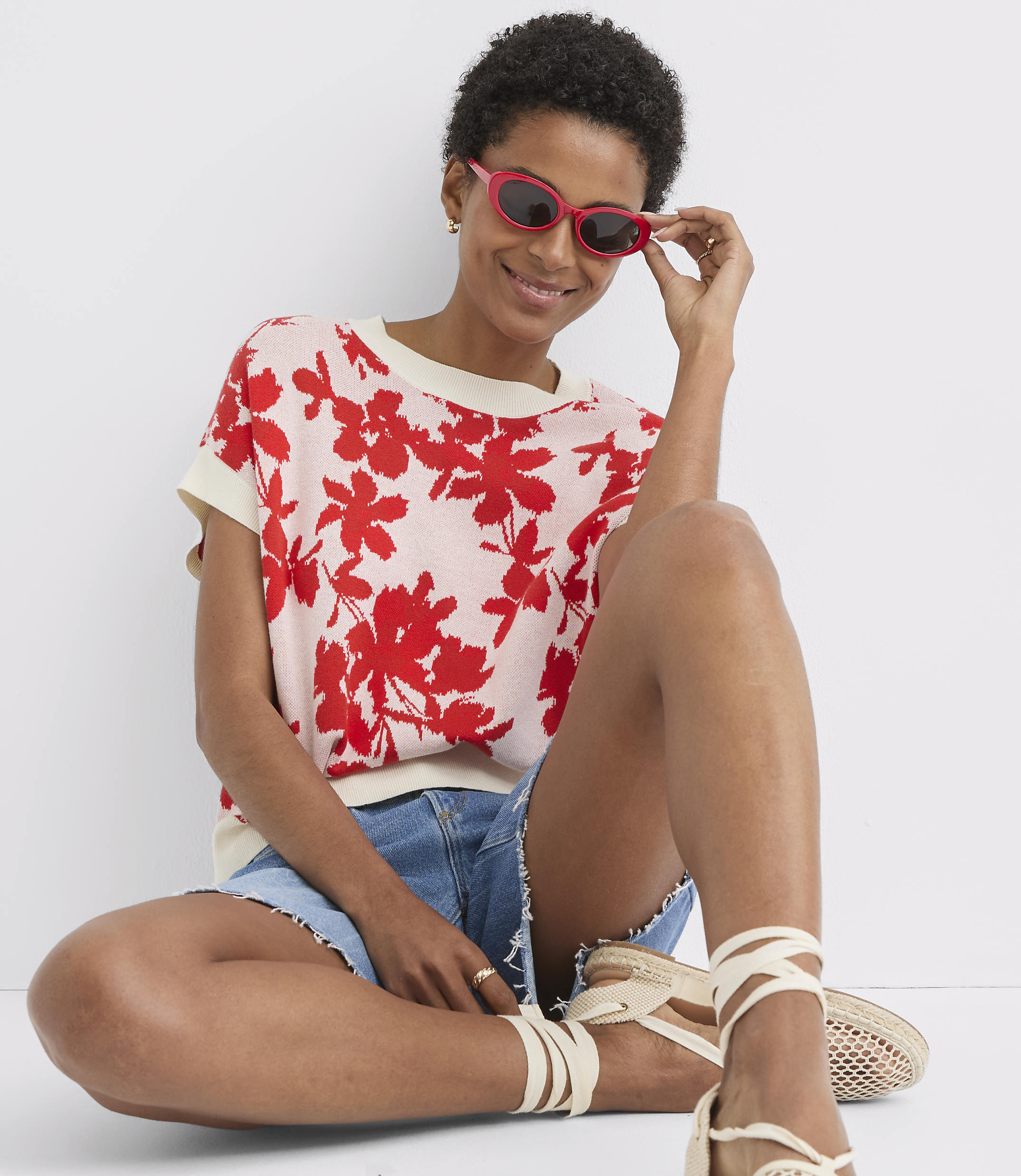 A woman in a floral top and denim shorts sits against a plain background, adjusting her pink sunglasses.