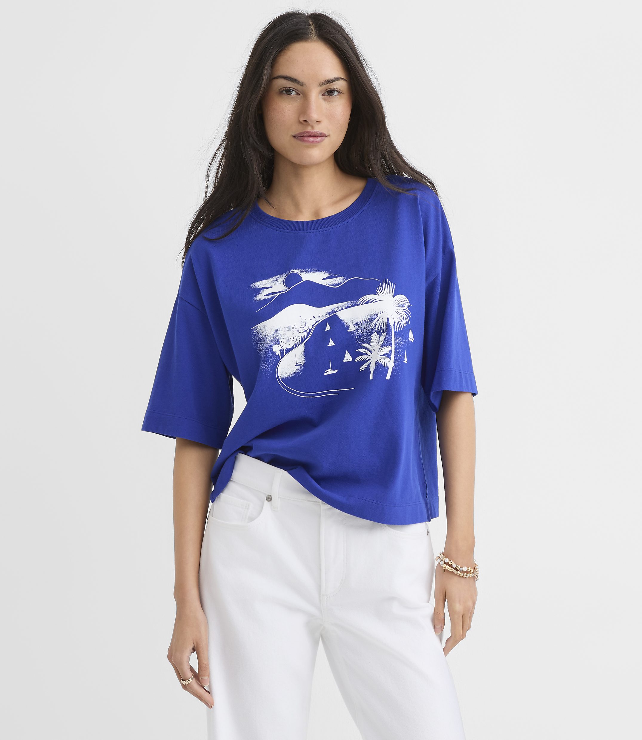 A woman in a blue t-shirt with a white graphic of a tropical scene featuring palm trees and sailboats stands against a plain background.