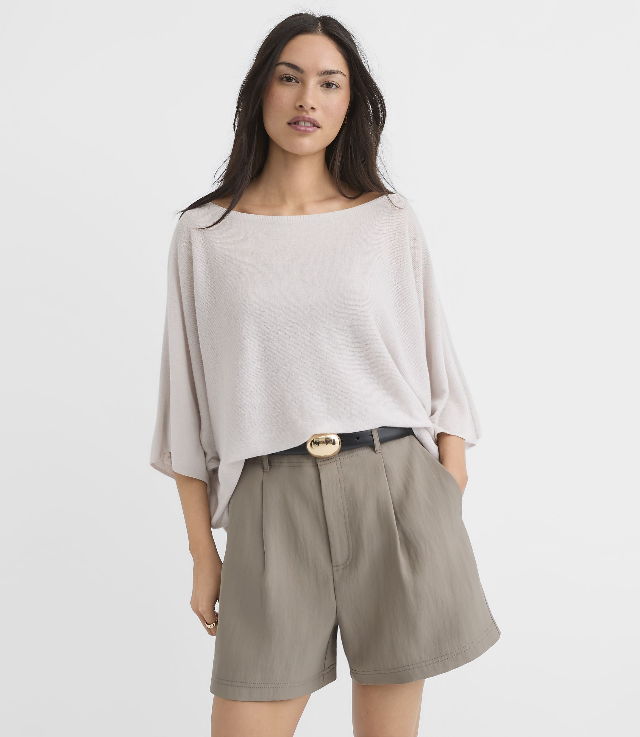 A woman in a light-colored oversized top and taupe shorts with a black belt stands against a plain white background.