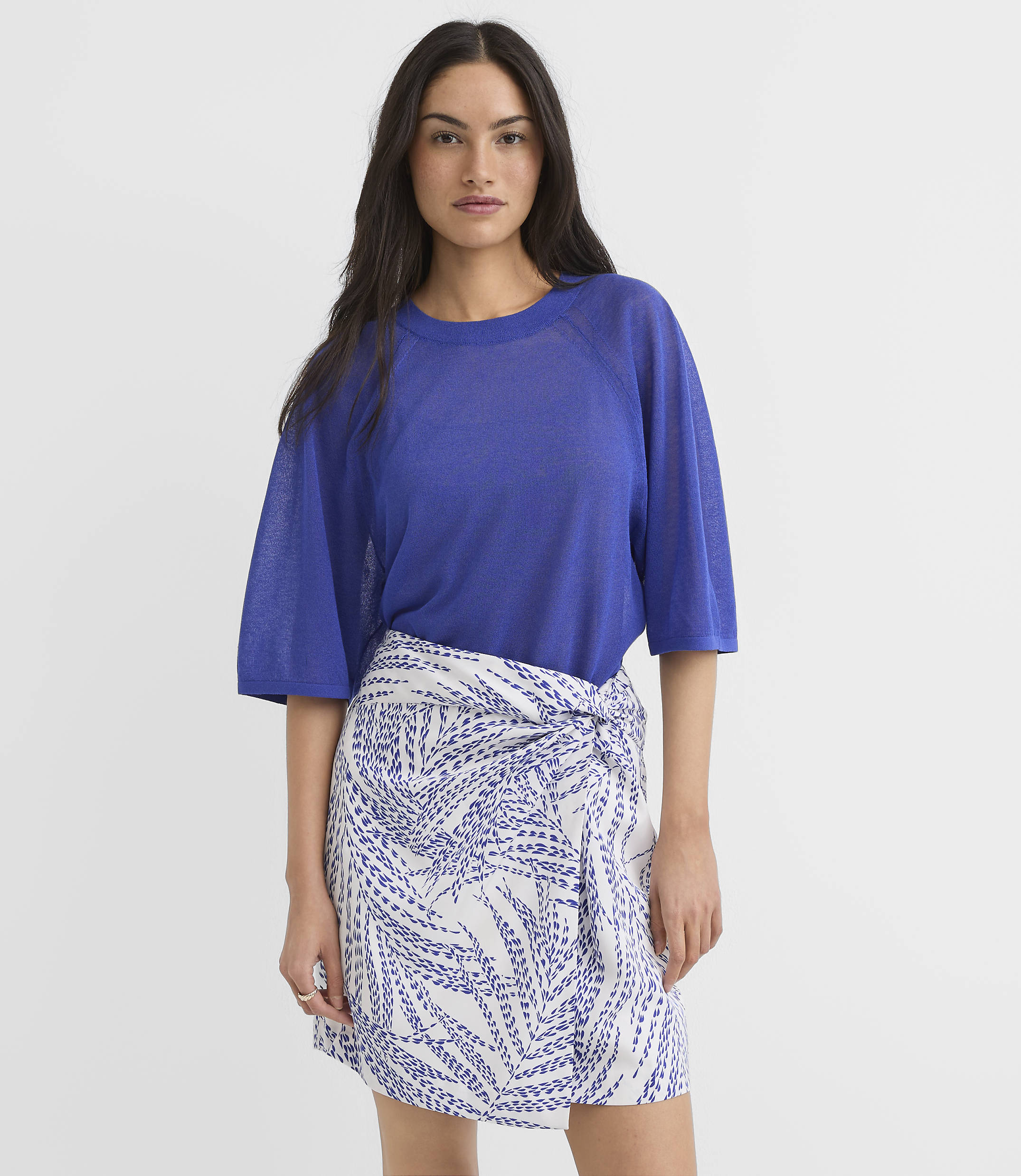 A woman in a blue top and a white and blue patterned skirt stands against a plain white background.