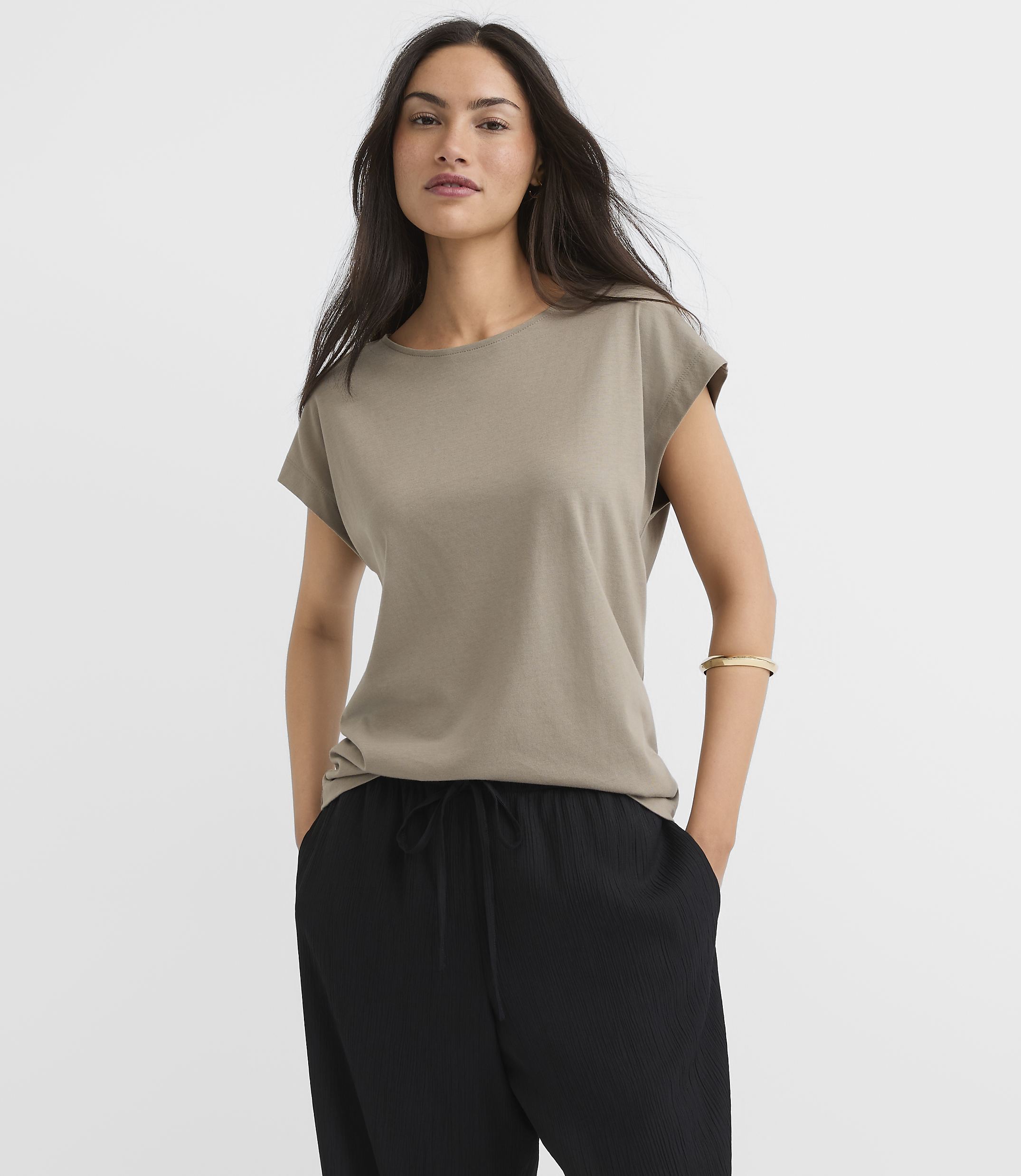 A woman in a taupe t-shirt and black pants stands against a plain gray background.