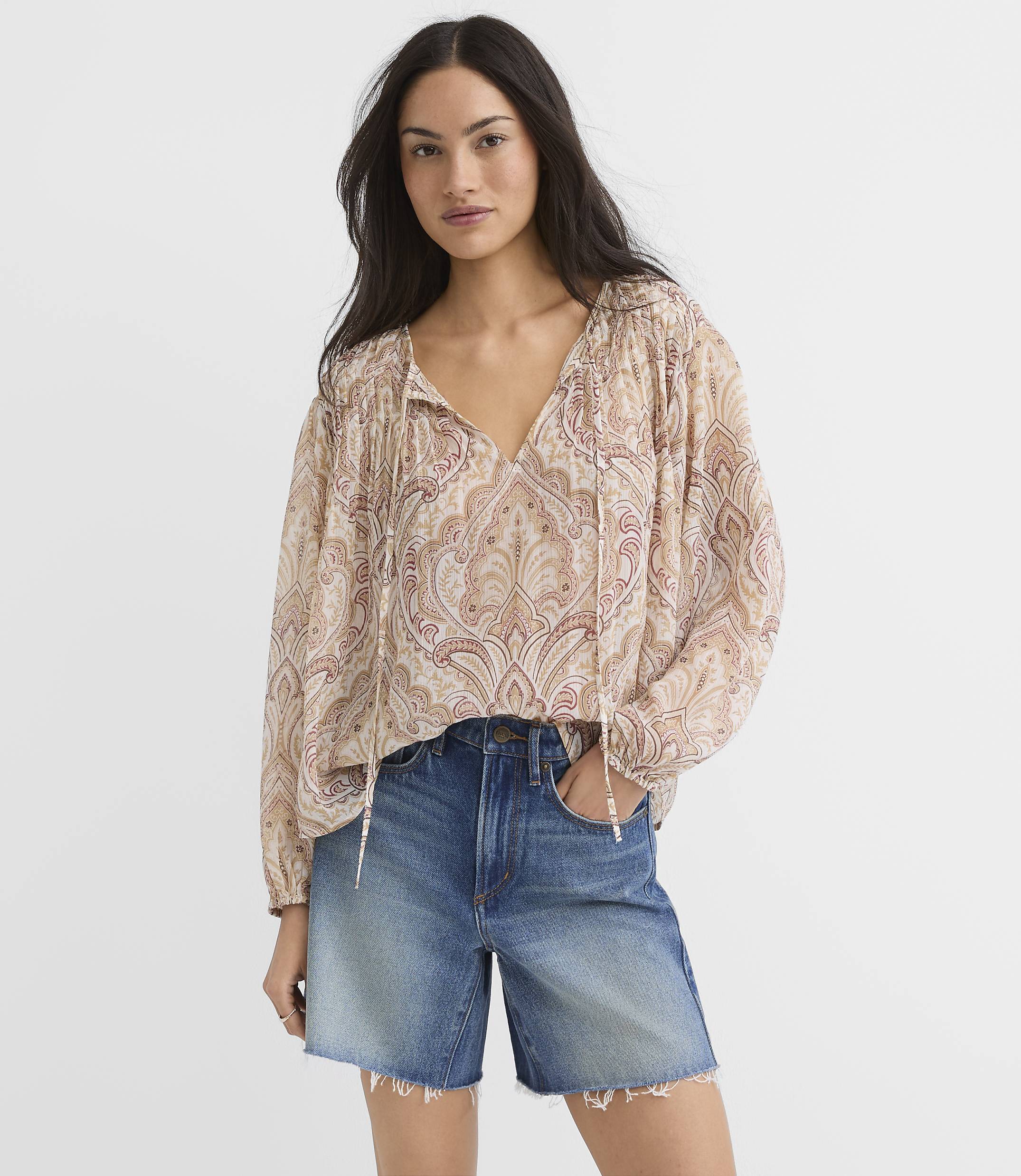 A woman in a patterned blouse and denim shorts stands against a plain white background.