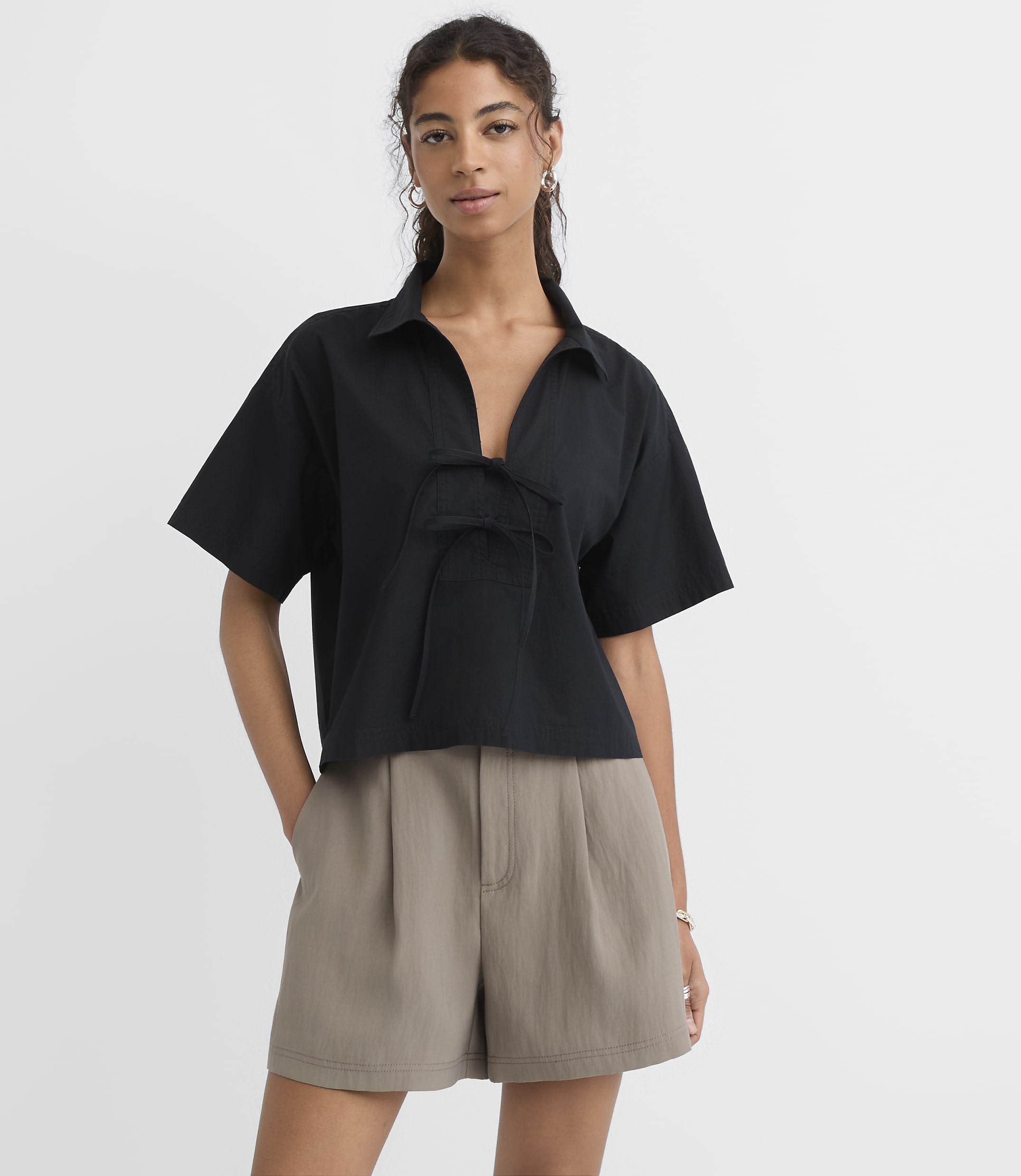 A woman in a black collared shirt and taupe shorts stands against a plain white background.