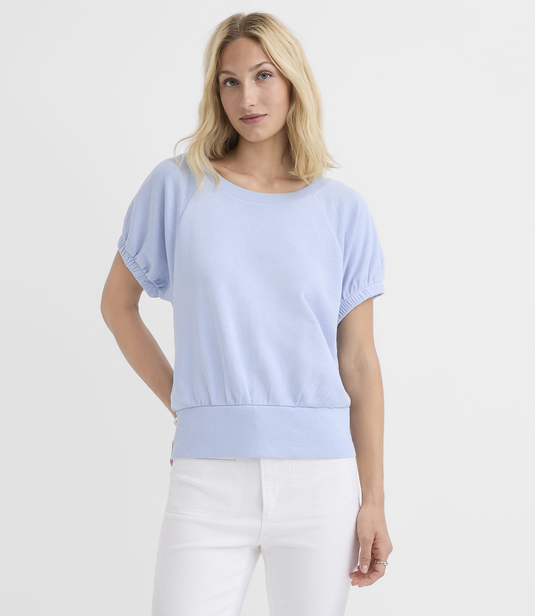 A blonde woman in a light blue top and white pants stands against a plain white background.