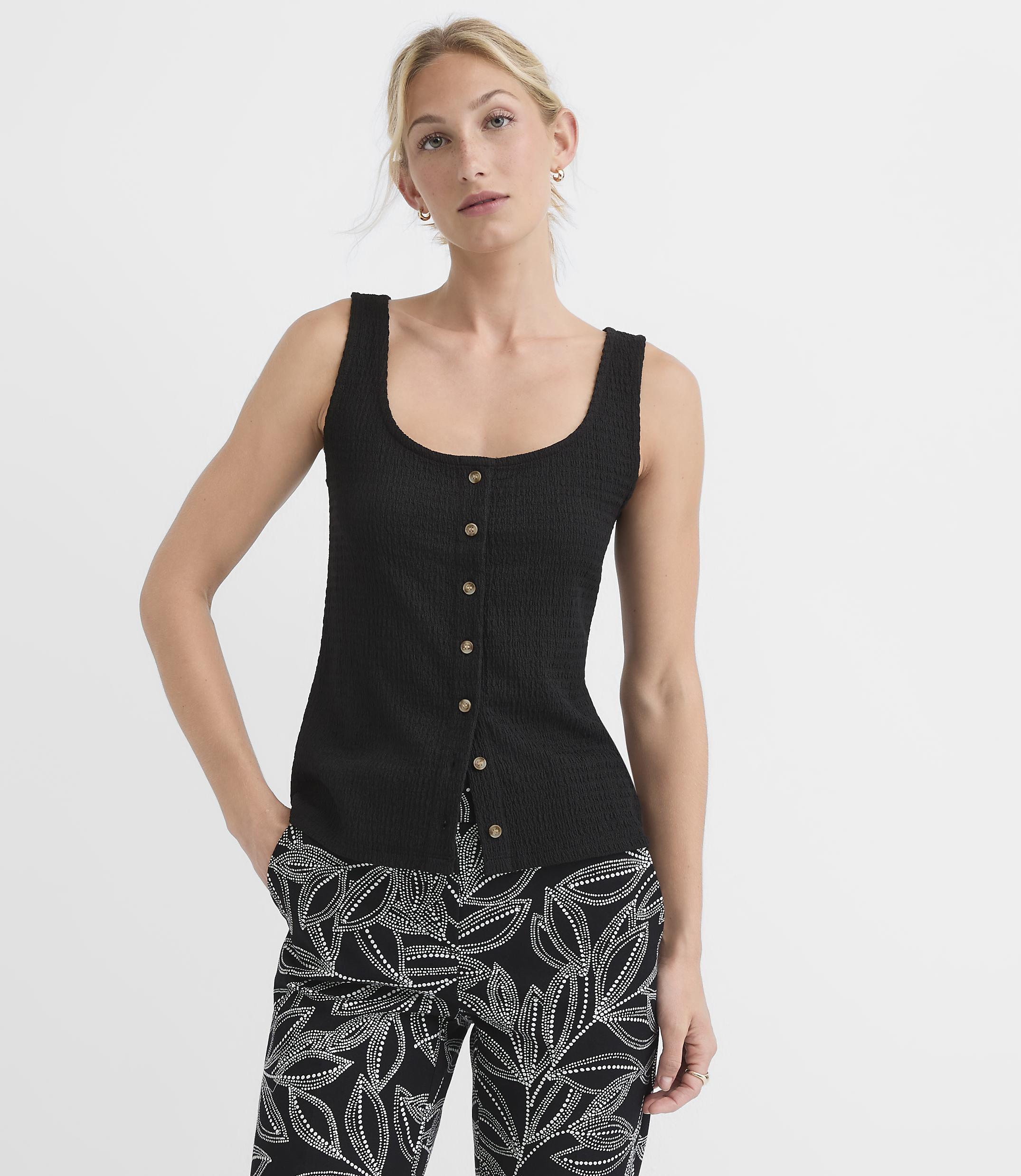 A woman in a black ribbed tank top and patterned pants stands against a plain white background.