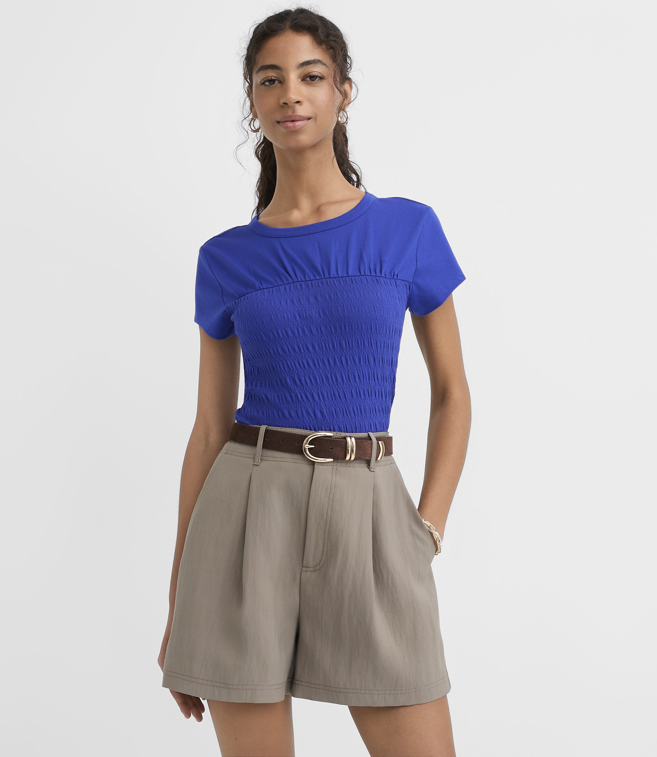 A woman in a blue smocked top and taupe shorts stands against a plain white background.