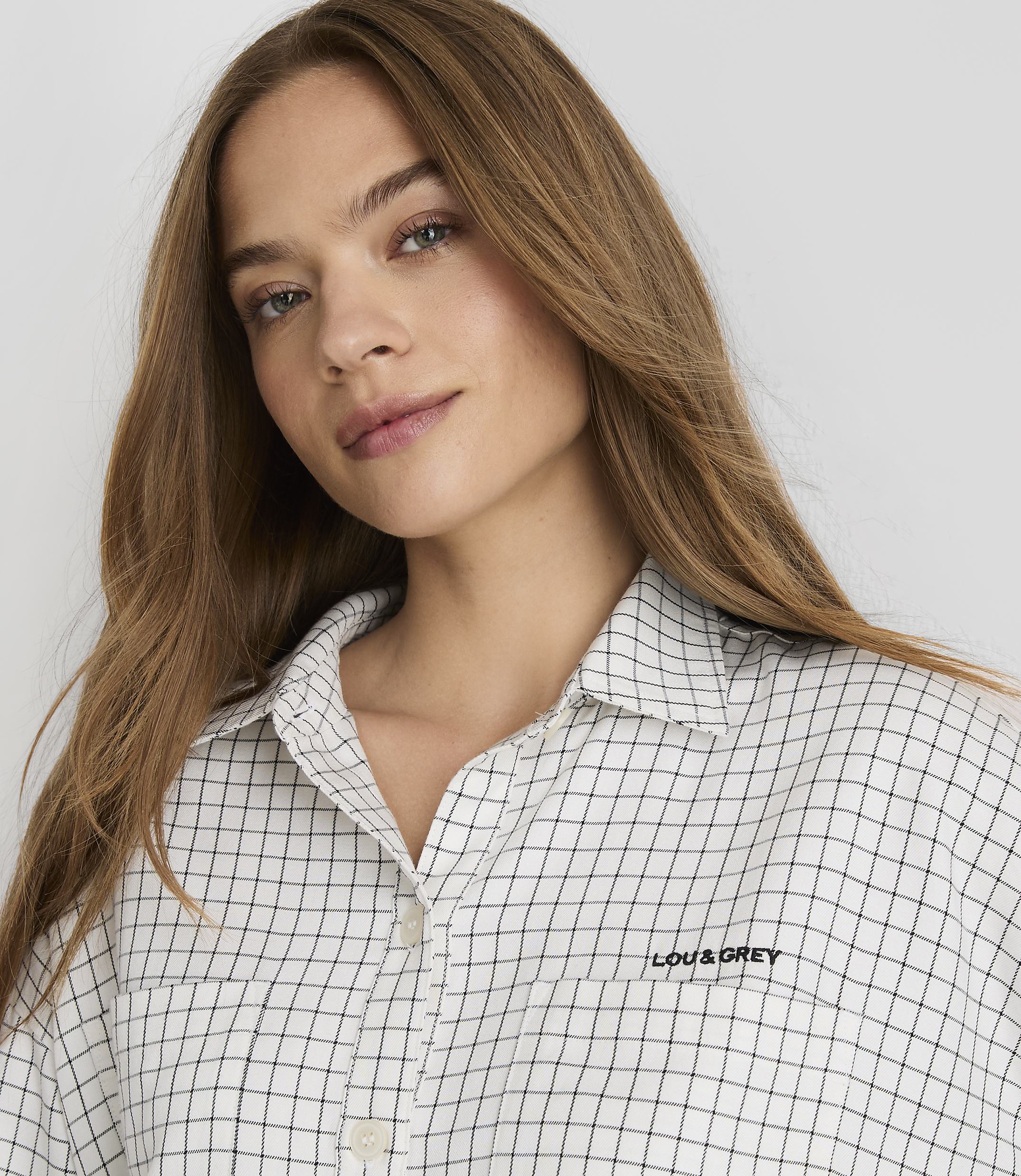 A young woman with long, light brown hair and a checkered shirt is looking forward against a plain white background.