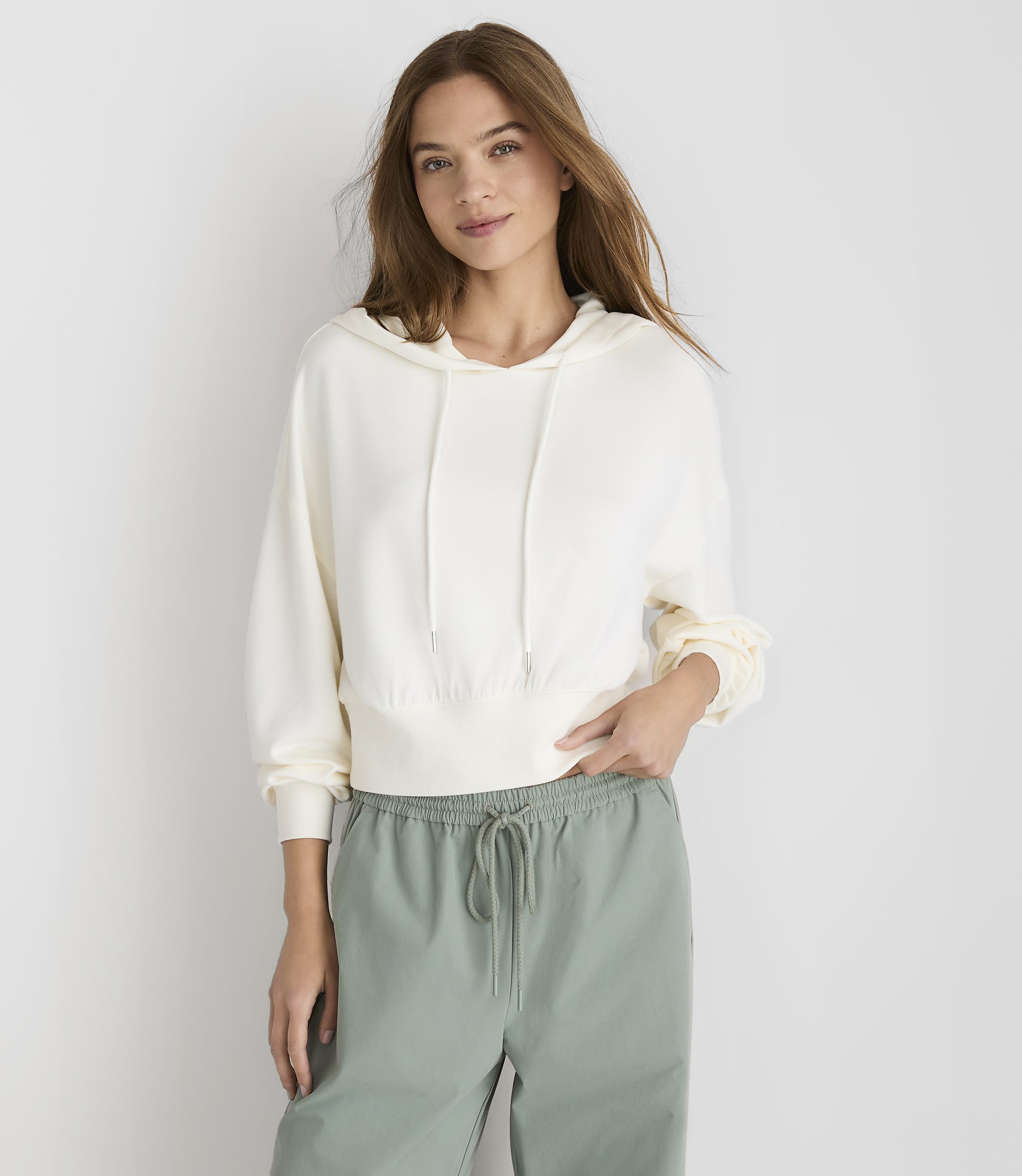 A young woman in a cream hoodie and green drawstring pants stands against a plain white background.