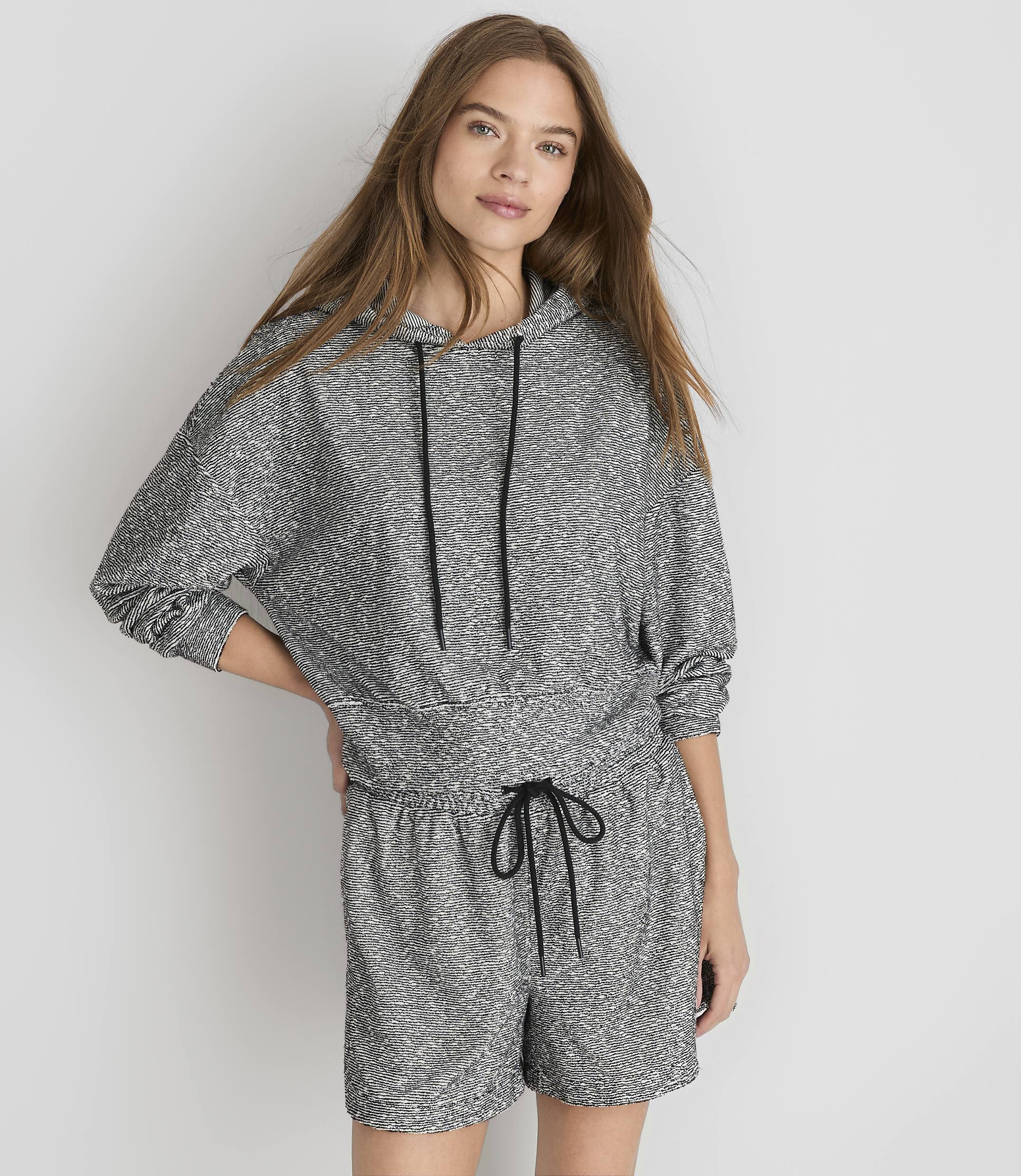 A young woman in a grey patterned hoodie and shorts stands against a plain white background.