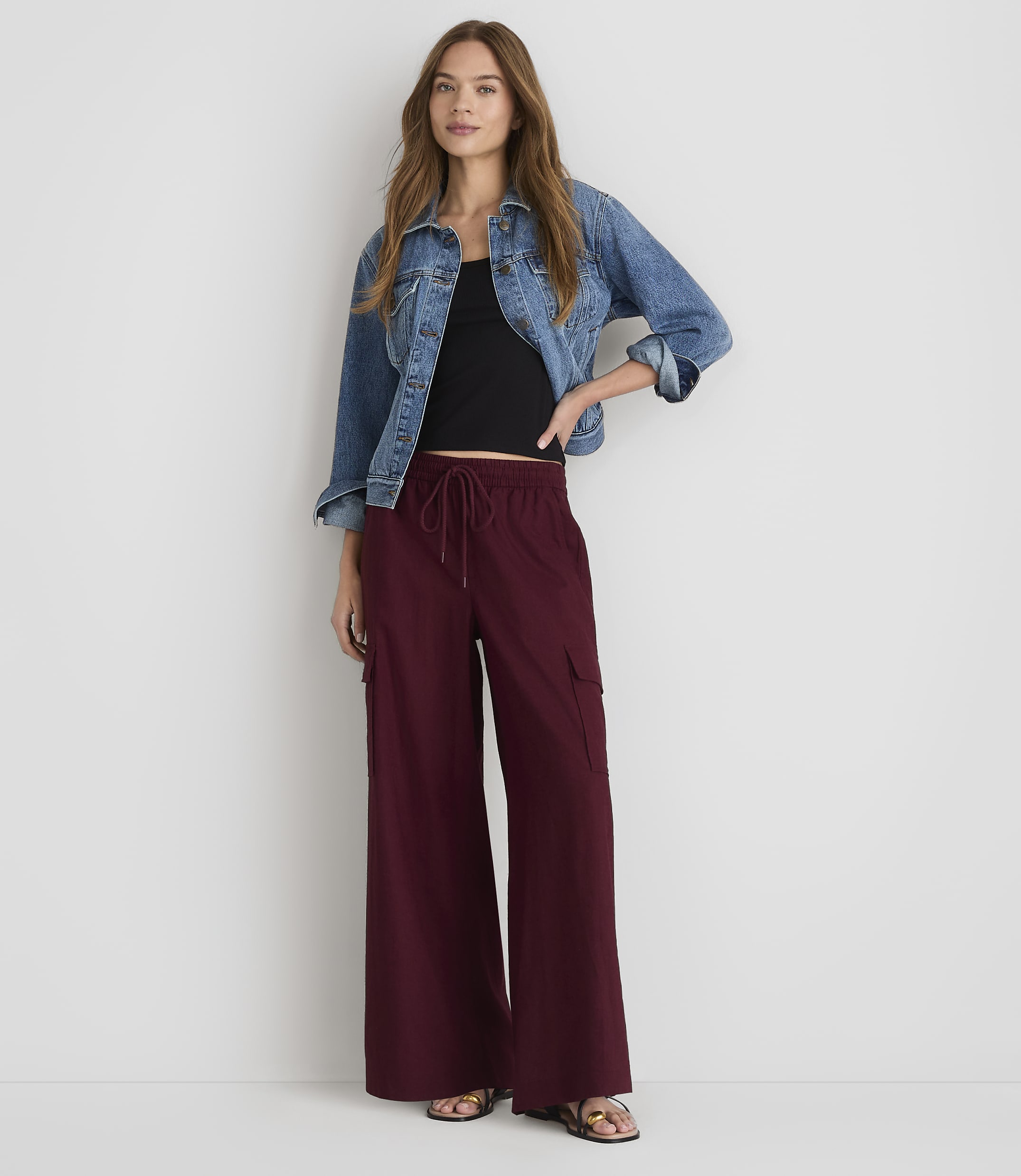 A woman in a denim jacket and maroon wide-leg pants stands against a plain white background.