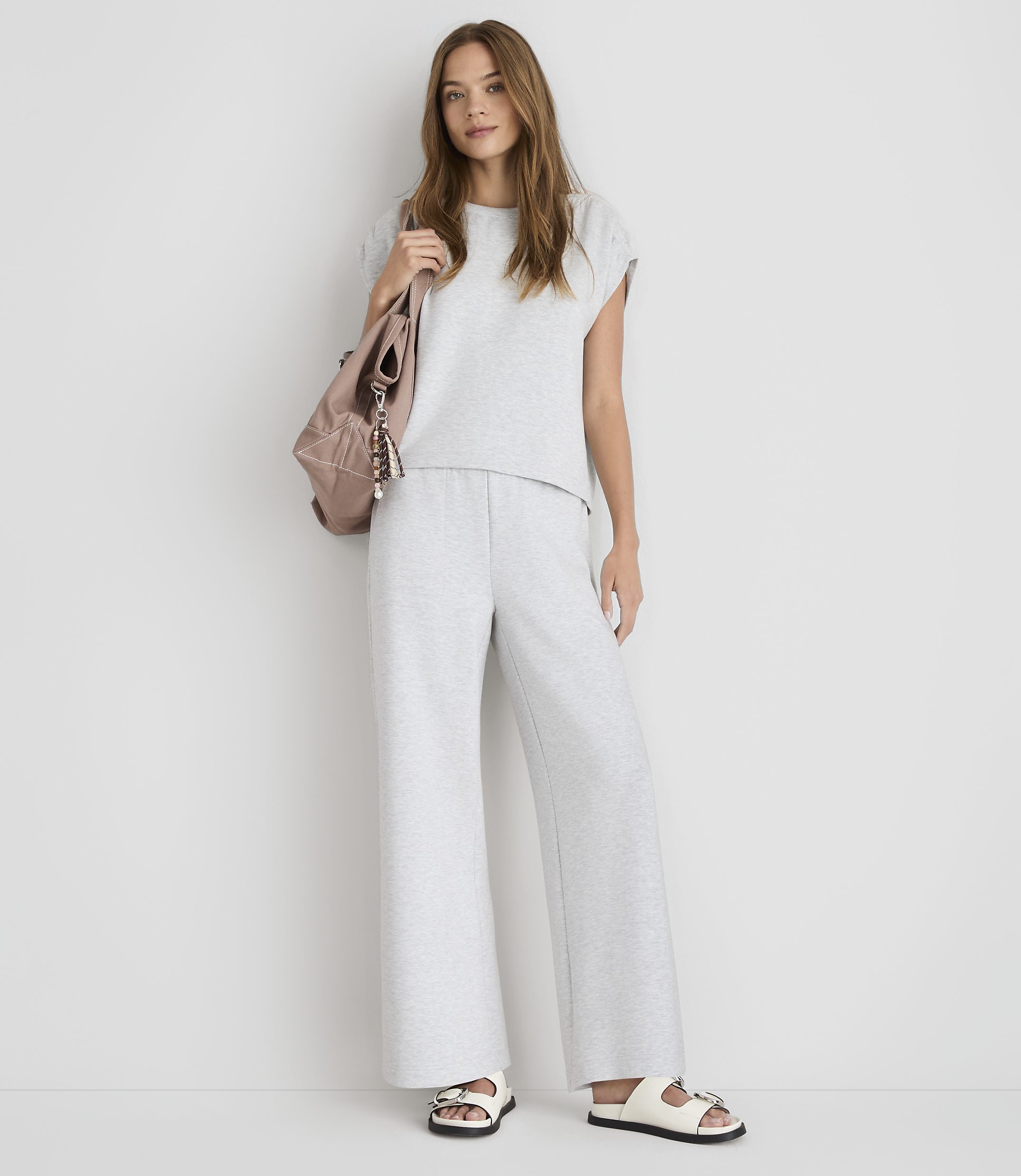 A young woman in a grey loungewear set and white sandals stands against a plain white background, with a pink backpack slung over her shoulder.