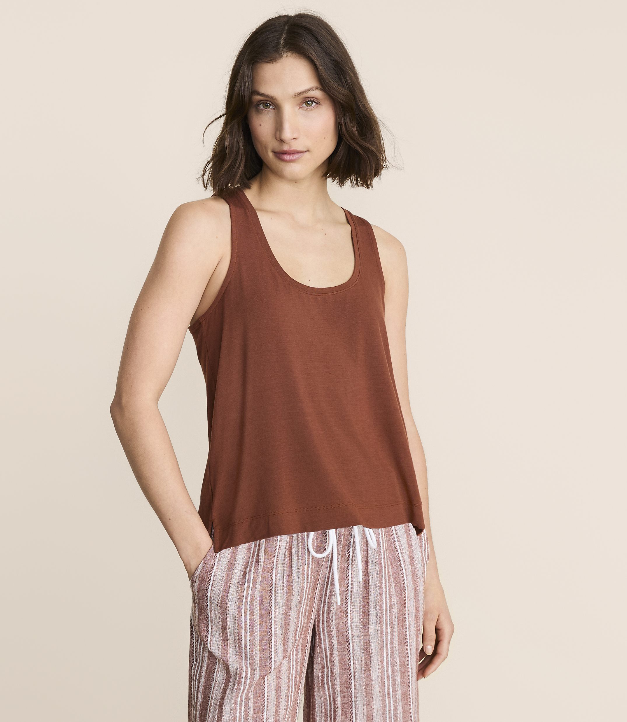A woman in a brown tank top and striped pants stands against a plain, light brown background.