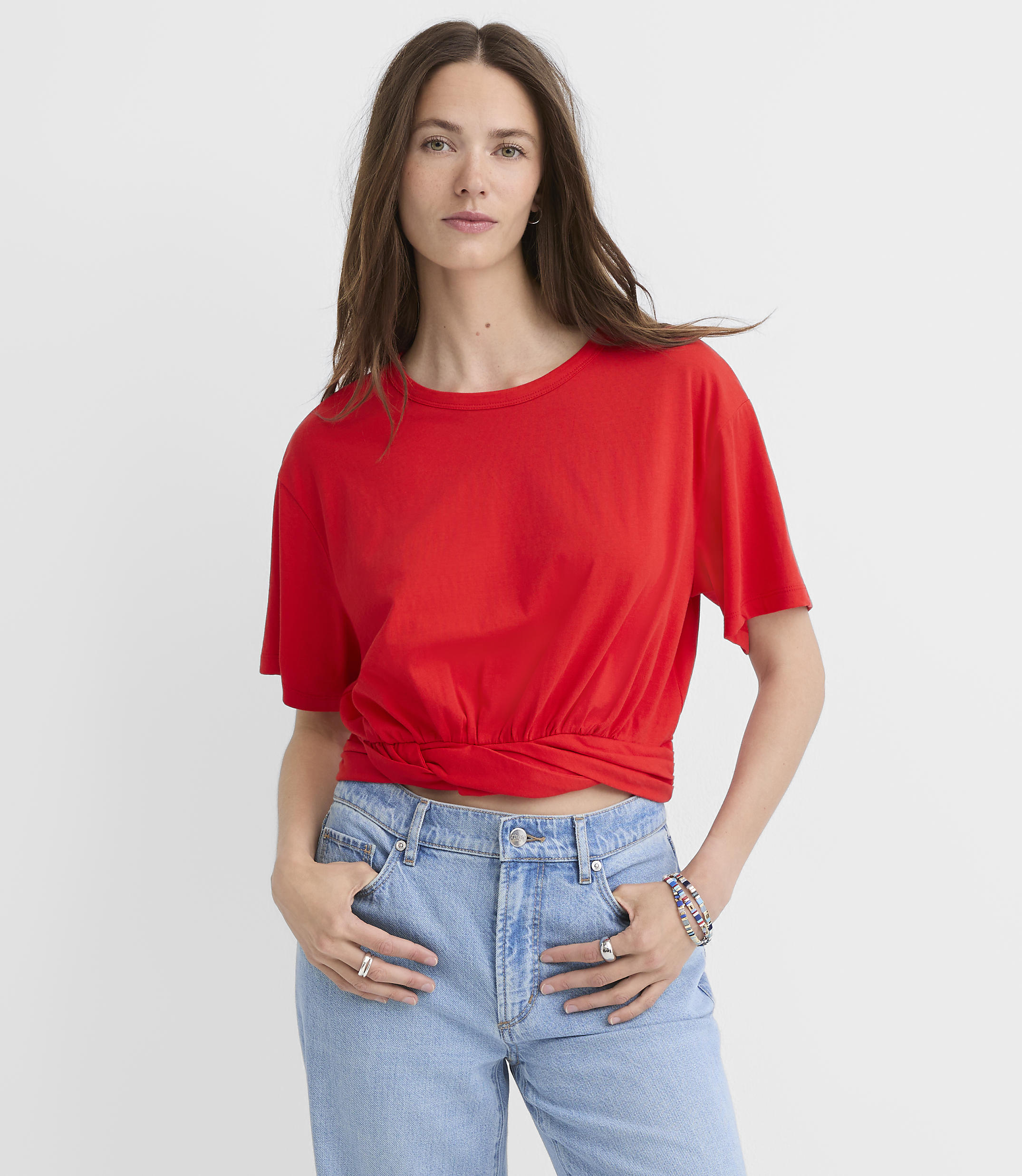 A woman in a red t-shirt and blue jeans stands against a plain white background.