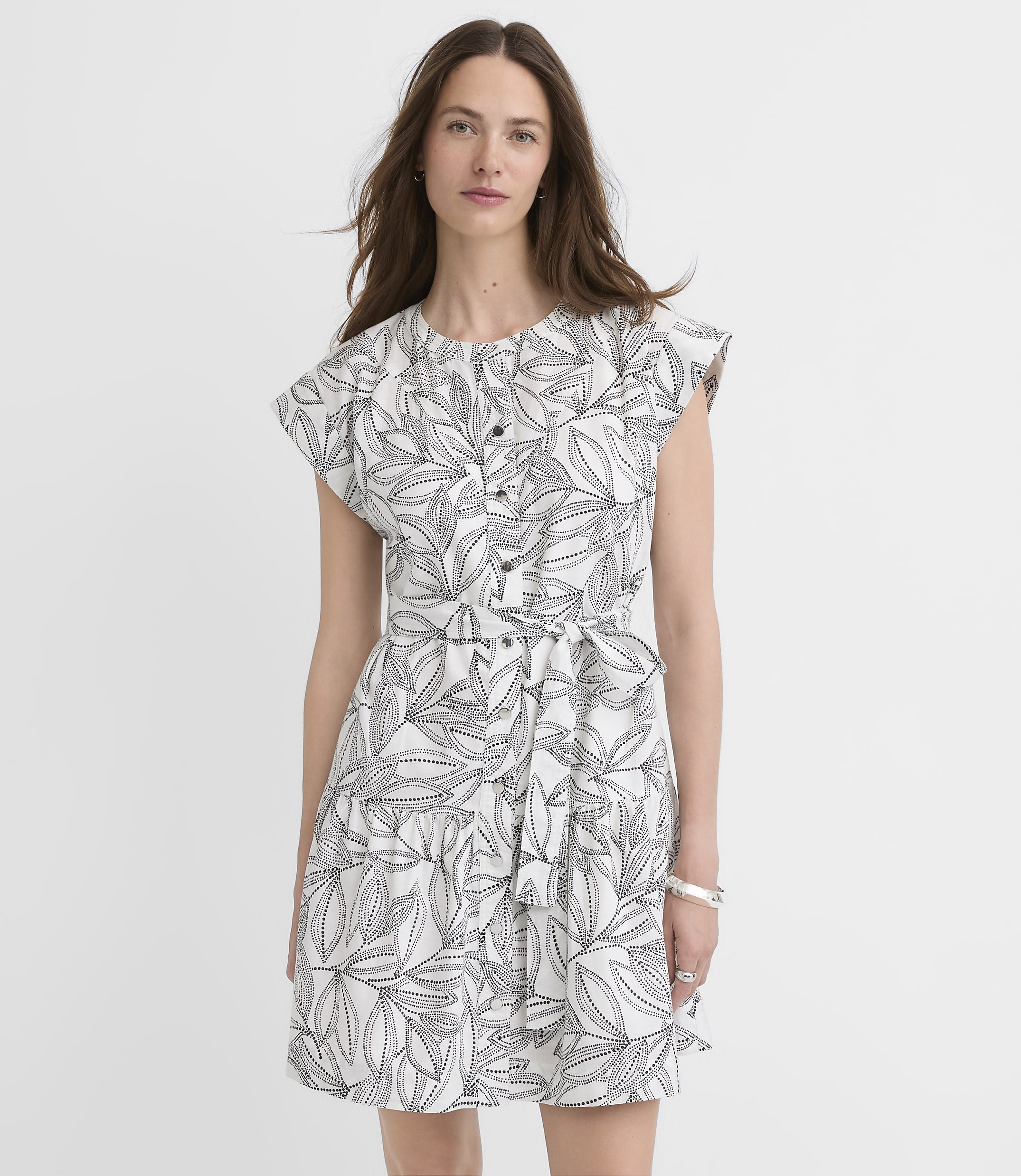 A woman in a white dress with a black leaf print stands against a plain white background.