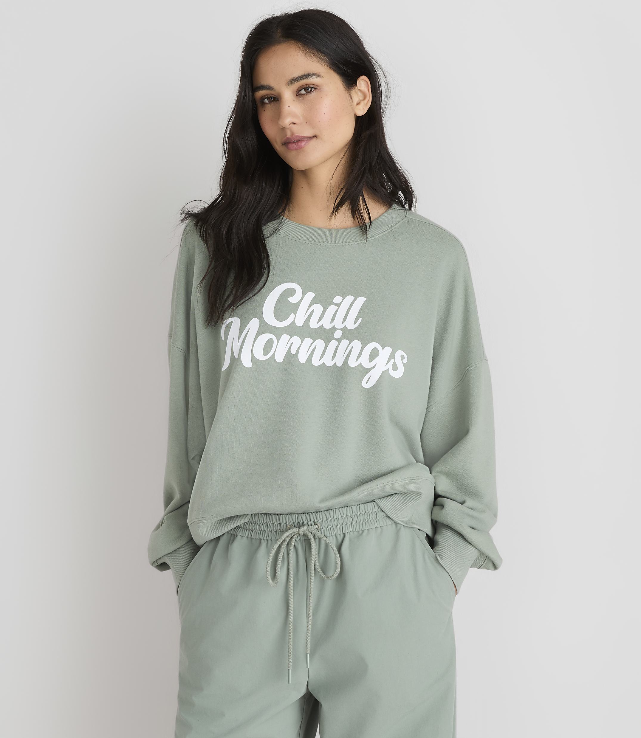 A woman in a light green sweatshirt with "Chill Mornings" printed on it stands against a plain white background.