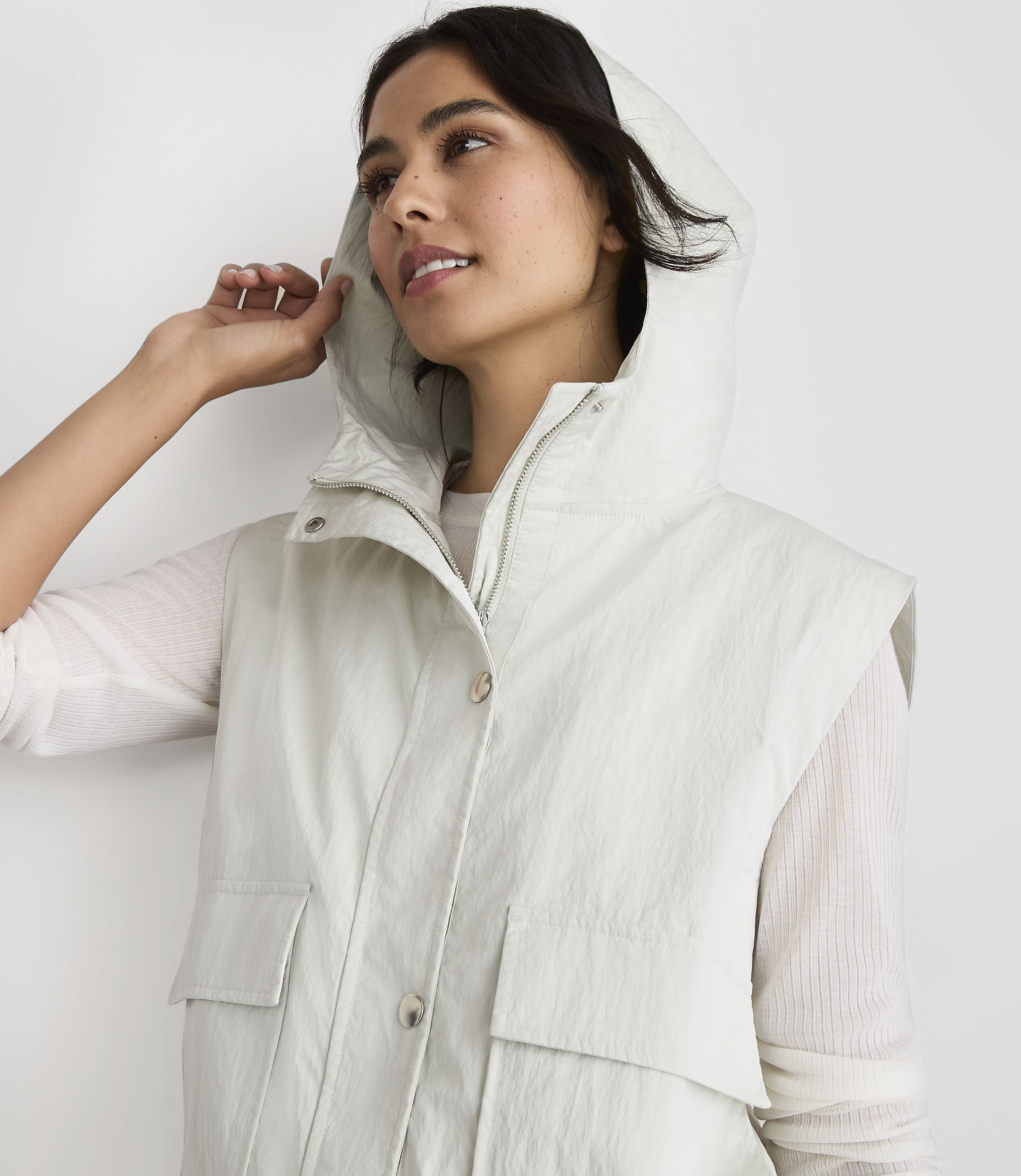 A woman in a light-colored hooded vest and white shirt looks to the side against a plain white background.