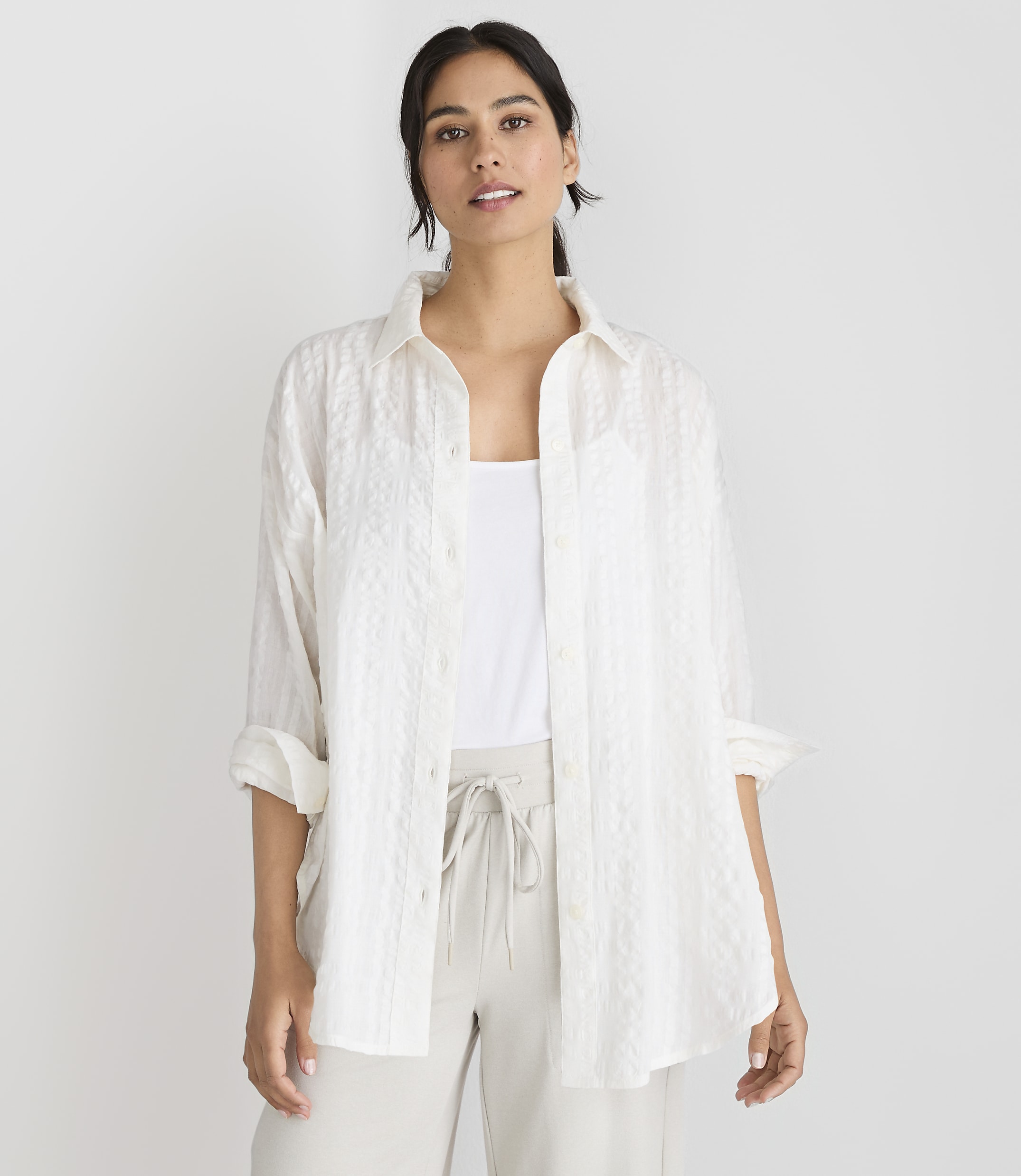 A woman in a white textured shirt and light-colored pants stands against a plain white background.