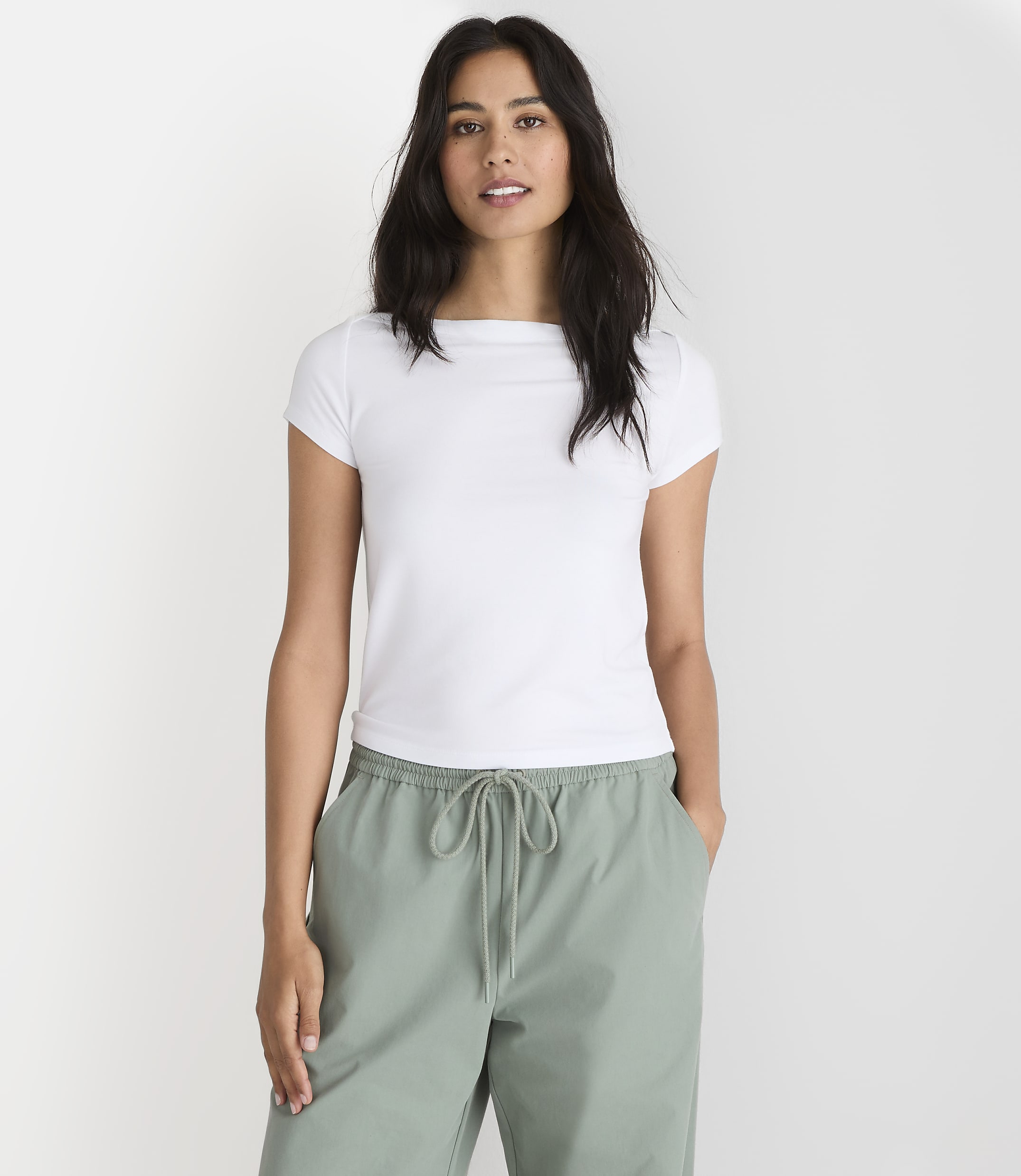 A woman in a white t-shirt and green pants stands against a plain white background.