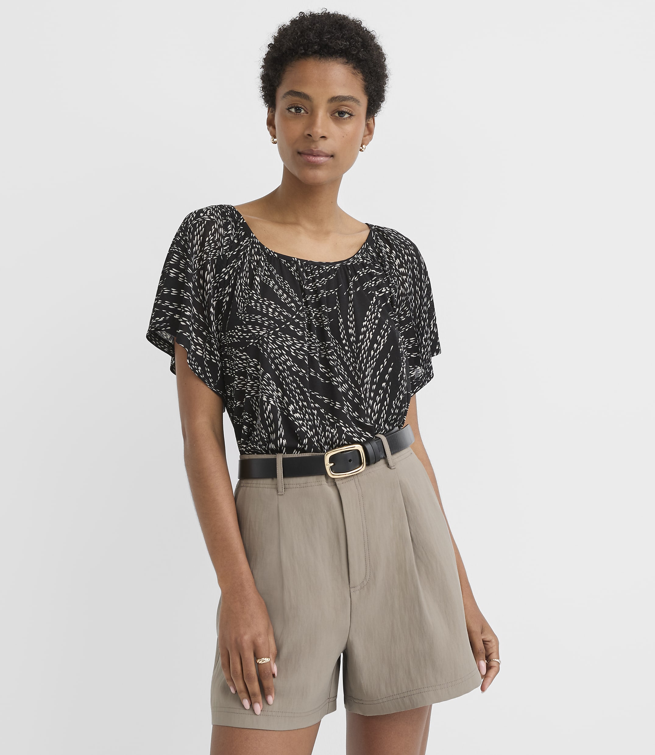 A woman in a patterned black top and taupe shorts stands against a plain white background.