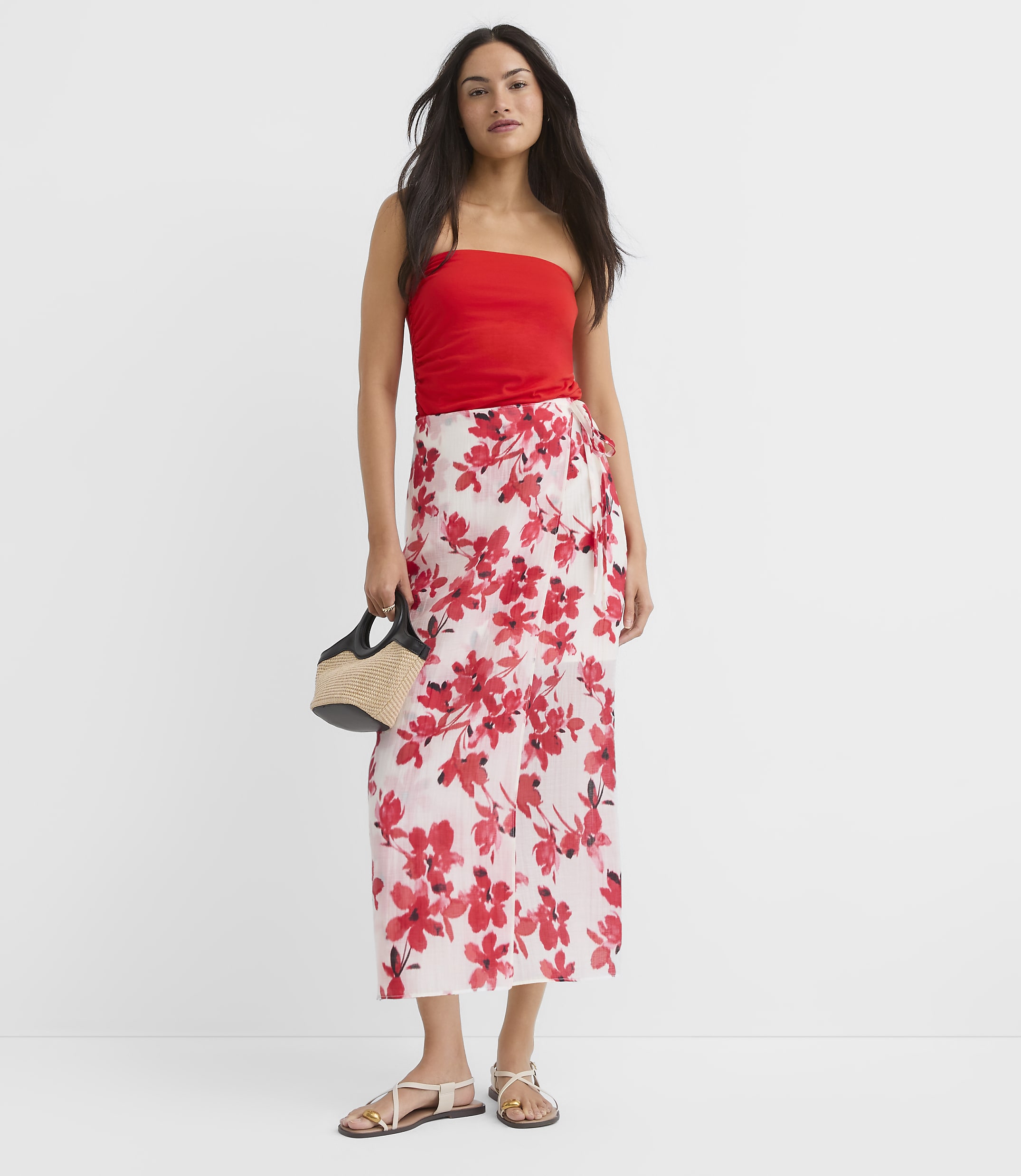 A woman in a red tube top and a floral wrap skirt stands against a plain white background, holding a straw bag.